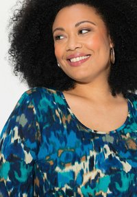 Sininen ja vihreä kuvioitu paita, jossa on pyöreä pääntie, esittää abstrakteja kuvioita turkoosin, ruskean ja vaalean kermaisen sävyissä. Sujuva kangastuntuma.