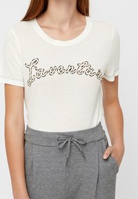 Witte T-shirt met korte mouwen met "Laventure" in een zwarte en gouden schreef gedrukt. Gecombineerd met grijze broek met trekkoord in de taille. Zachte stoftextuur.