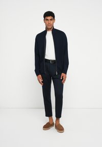 Casaco azul-marinho com fecho zip, gola alta, sobre uma camisa branca, combinado com calças escuras e sapatos castanhos de slip-on. Design minimalista e ajuste sob medida.
