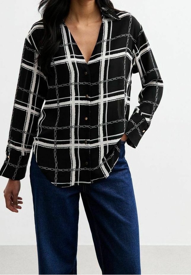 Femme portant une chemise noire à manches longues boutonnée avec un motif de chaînes et de grille blanche, associée à un jean bleu foncé, main dans la poche.