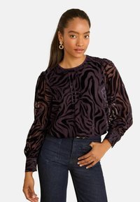 Blusa nera trasparente con stampa zebra viola, dettaglio plissettato sul davanti, scollatura rotonda e maniche a sbuffo. Abbinata a jeans in denim scuro.