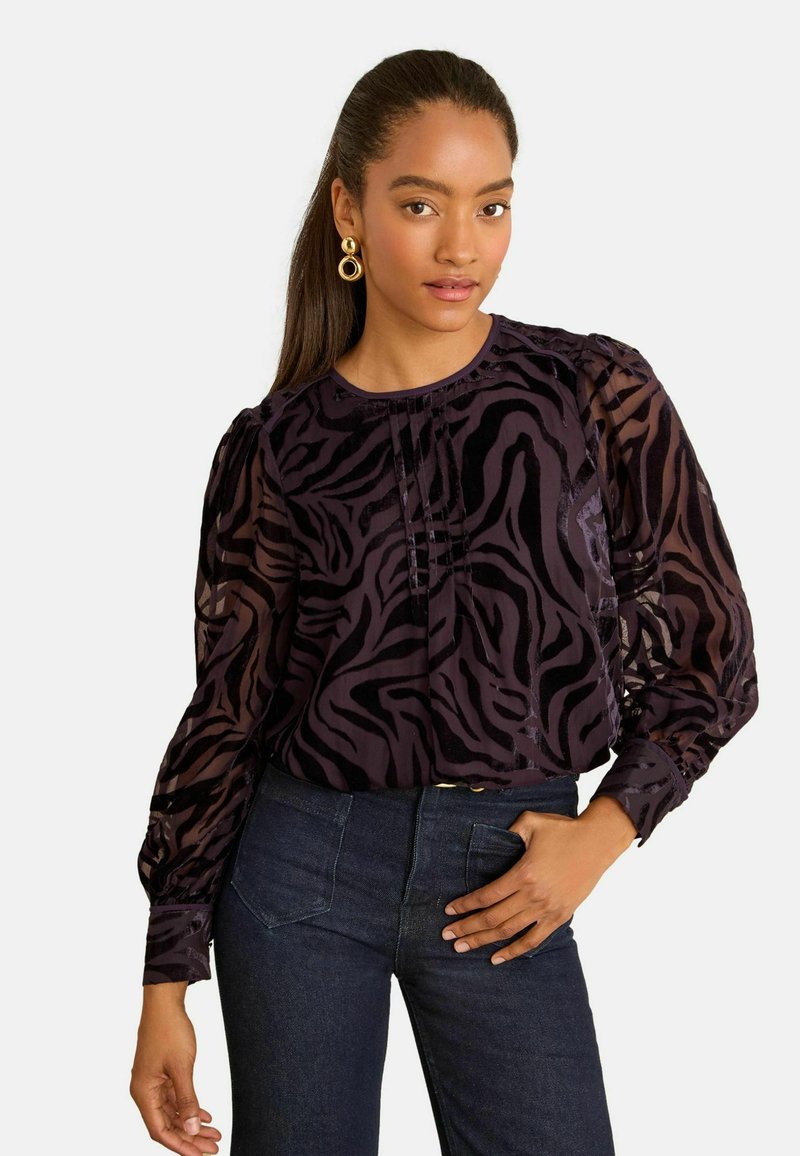 Blusa nera trasparente con stampa zebra viola, dettaglio plissettato sul davanti, scollatura rotonda e maniche a sbuffo. Abbinata a jeans in denim scuro.
