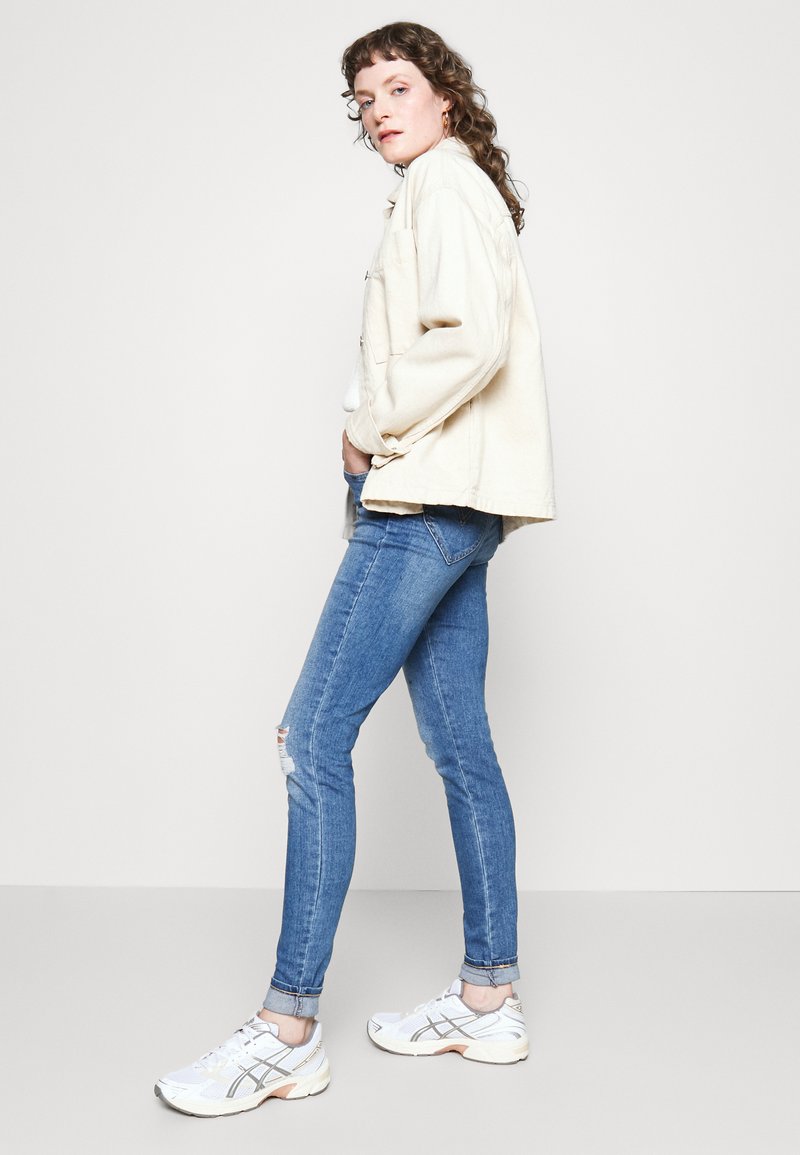 Giacca di jeans beige dal fit rilassato, abbinata a jeans skinny azzurri chiari con strappo e scarpe sportive bianche con dettagli grigi.