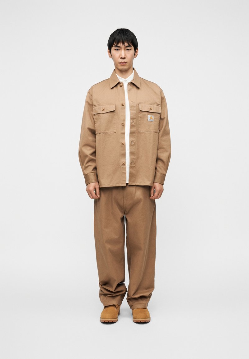 Tan cotton shirt jacket with front pockets, button closure, and logo patch, paired with matching loose-fit pants and light brown footwear.