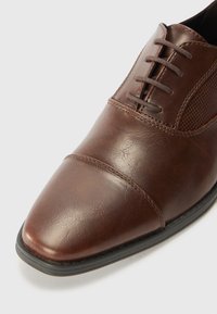 Chaussure en cuir marron avec un bout pointu, un laçage, un panneau latéral texturé et des coutures contrastées sur une semelle en caoutchouc noire.