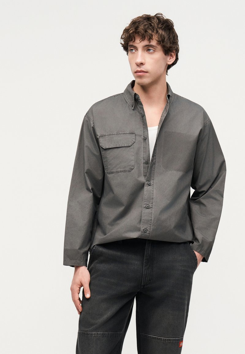 Giovane uomo con capelli ricci che indossa una camicia grigia abbottonata sopra una maglietta bianca e jeans scuri, che guarda verso sinistra su uno sfondo chiaro.