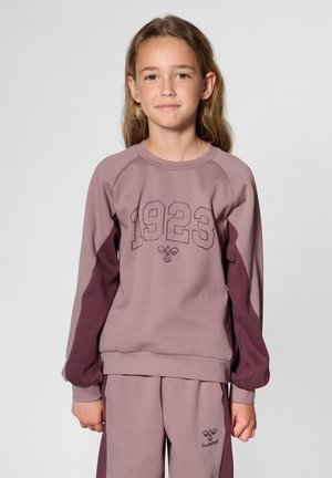 Ung pige står med fronten mod kameraet iført en sart lilla og mørk lilla sweatshirt og bukser med "1923" og bi-logo på forsiden.