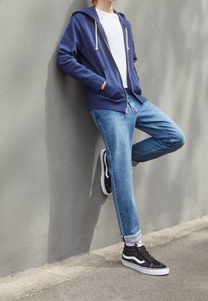 Person trägt einen blauen Hoodie, weißes T-Shirt, hochgekrempelte Jeans und schwarze High-Top-Sneaker und lehnt draußen an einer grauen Wand.