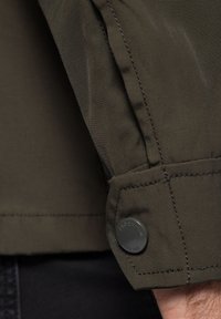 Ciemnozielona kurtka wykonana z teksturowanej tkaniny, z zapięciem na guziki przy mankietach i widocznym logo. Projekt charakteryzuje się czystym, nowoczesnym krojem oraz widocznymi szwami.