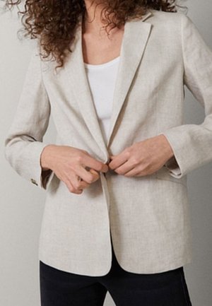 Kvinde med krøllet hår, der knapper en lys beige blazer over en hvid skjorte, iført sorte bukser foran en ensfarvet baggrund.