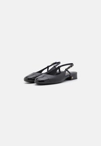 Czarne buty na niskim obcasie typu slingback z błyszczącymi czubkami i regulowanymi paskami z klamrami na jednolitym jasnym tle.