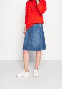 Gonna di jeans con taglio a trapezio, di lunghezza media, caratterizzata da grandi bottoni dorati sul davanti, abbinata a una felpa con cappuccio rossa brillante e sneakers bianche.