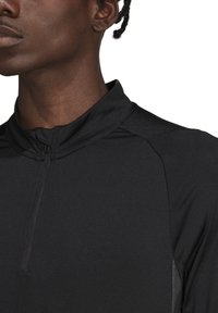 Top sportivo nero con colletto alto e zip, realizzato in materiale liscio. Presenta dettagli riflettenti lungo i lati per aumentare la visibilità.