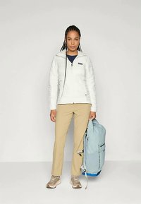 Bela izolirana jakna z zadrgo, bež hlače in svetlo modra nahrbtnik. Obut tan športni copati, stoji pred nevtralno ozadje.