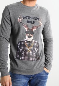 Felpa grigia con una grafica di un cervo con corna che indossa occhiali da sole, sovrapposta a una camicia a quadri.