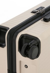 Valigia rigida beige con superficie testurizzata, zip nera e ruote girevoli doppie; presenta una maniglia laterale e un meccanismo di chiusura sicuro.