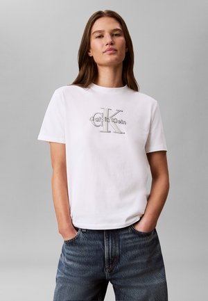 Giovane donna con lunghi capelli castani che indossa una t-shirt bianca Calvin Klein e jeans blu, in piedi con le mani nelle tasche davanti a uno sfondo semplice.