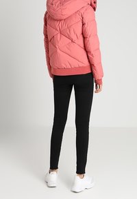 Doudoune rose matelassée en forme de losanges, dotée d'une capuche et de poignets côtelés, portée sur un jean skinny noir et des baskets blanches.