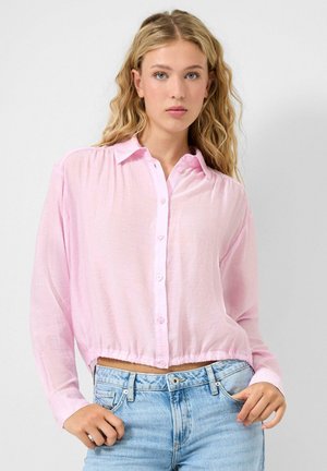 Junge Frau mit welligem blondem Haar, die eine hellrosa Bluse mit Knöpfen und hellblaue Jeans trägt, vor einem schlichten weißen Hintergrund stehend.