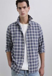 Camisa de cuadros en marinero y blanca de manga larga, hecha de algodón suave, con cuello abotonado y mangas arremangadas. Llevada sobre una camiseta blanca.