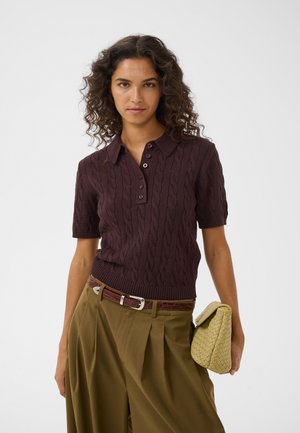 Frau mit lockigem Haar, die ein dunkelbraunes kurzärmeliges Poloshirt mit Zopfmuster, olivfarbene weit geschnittene Hose, braunen Gürtel trägt und eine gewebte beige Clutch hält.