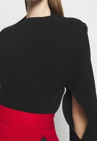 Schwarzes geripptes Strickoberteil mit hochgeschlossenem Kragen und ausgeschnittenen Ärmeln, kombiniert mit einem roten Taillengürtel, der mit Knopfakzenten versehen ist.