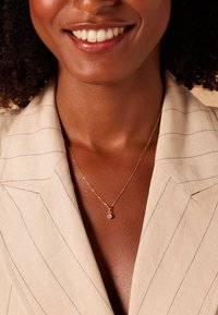 Collier en chaîne dorée avec un petit pendentif en gemme claire, porté sur un blazer beige clair à rayures avec une texture douce.