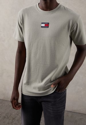 Man draagt een lichtgrijs Tommy Jeans T-shirt en donkere spijkerbroek, staat met één hand in de zak tegen een neutrale achtergrond.