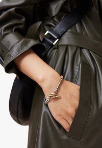 Bracciale in catena d'oro e argento con chiusura a moschettone, indossato su una mano. Sullo sfondo, un cappotto in pelle verde oliva con maniche ripiegate.