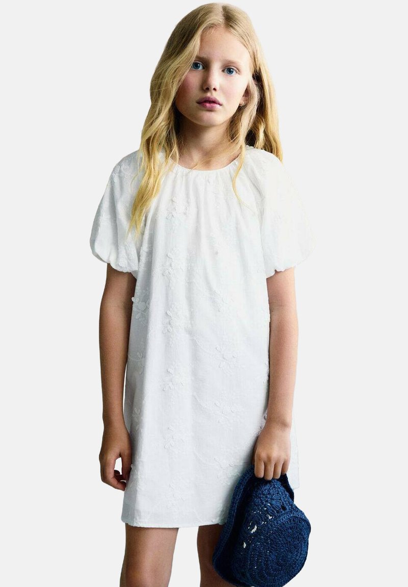 Vit kort klänning med puffärmar som har blommönsterbroderi. Modellen håller en blå vävd hatt som ger kontrast till outfiten.
