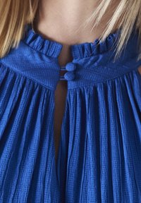 Robe bleue texturée avec un col froncé et une ouverture en forme de trou de serrure, dotée de deux boutons ronds au niveau du cou, et de plis verticaux.