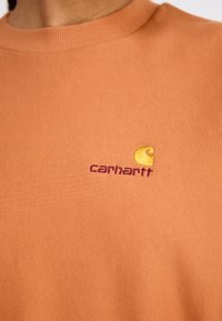 Orange sweatshirt lavet af blødt stof med ribbet rund hals. Broderet Carhartt-logo i lilla og gul på brystet.