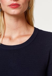 Kvinna i en ribbad, mörkblå tröja med rund halsringning och långt, rakt blont hår, där hon bär tunna svarta och silvriga hängande örhängen.