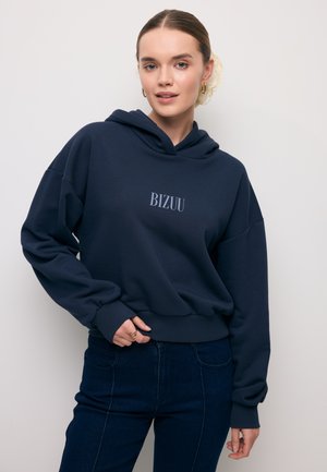Jonge vrouw draagt een marineblauwe hoodie met de tekst "BIZUU", een blauwe spijkerbroek, gouden oorringen en een beige haaraccessoire, staand tegen een witte muur.