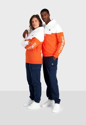 Deux individus portent des sweat-shirts assortis avec un design orange et blanc ; tous deux portent des pantalons bleu marine. Des baskets blanches complètent leurs tenues.