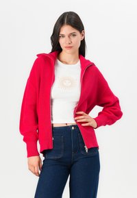 Fuchsia geribbelde ritstrui met hoge kraag, met brede mouwen en textuurbreiwerk. Gepaard met een witte blouse en high-waisted jeans.