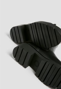 Semelle de botte en cuir noir avec un caoutchouc noir épais et rainuré. Présente un motif texturé pour l'adhérence et des bords arrondis pour le confort.