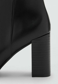 Črni usnjeni gležnjar z teksturirano površino, s kvadratnim, robustnim peto in gladkim, elegantnim dizajnom. Na strani je viden šiv.