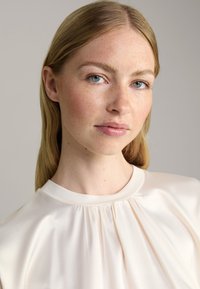 Kurzärmlige Seidenbluse in zartem cremigen Farbton, mit plissiertem Ausschnitt und glatter Textur, rundem Kragen und minimalistischem Design.