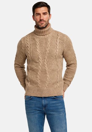 Homme portant un pull à col roulé beige en tricot torsadé et un jean bleu, debout devant un fond clair uni, les mains dans le dos.