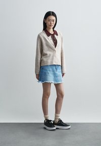 Cardigan beige clair à boutons sur une chemise col maroon, associé à une jupe en denim bleu clair effilochée et des baskets noires.