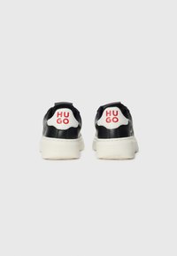 Des baskets noir et blanc vues de dos, avec des semelles blanches épaisses et "HUGO" en rouge sur les languettes blanches du talon.