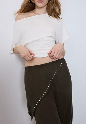 Frau trägt ein weißes, schulterfreies Oberteil und einen dunklen asymmetrischen Rock mit metallischen Nieten, Hände justieren das Oberteil in Taillennähe.