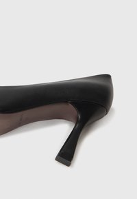 Chaussure à talon haut en cuir noir avec un bout pointu, surface texturée et un talon fin. Pas d'embellissements ou de motifs visibles.
