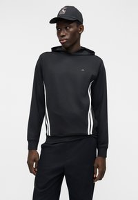 Sweatshirt preta com capuz e riscas brancas laterais, feita de um tecido liso. Combinada com calças pretas e um boné a condizer.