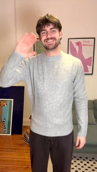 Χαμογελαστός άνδρας με γκρι πουλόβερ χαιρετά με το δεξί χέρι σε σαλόνι με έργα τέχνης ζώων και πόστερ με την επιγραφή "ΣΤΑΜΑΤΗΣΤΕ ΤΟΝ ΠΑΝΙΚΟ" στο φόντο.