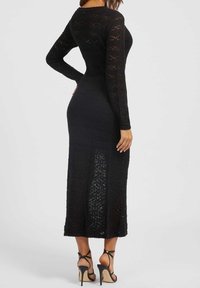 Robe noire en maille à manches longues avec une silhouette ajustée, un motif complexe en dentelle et un ourlet ouvert, associée à des talons hauts noirs à lanières.