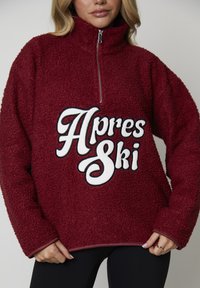 Burgundowy polarowy sweter z wysokim kołnierzem i pół zamkiem. Z przodu znajduje się biały, haftowany napis "Apres Ski". Miękki, strukturalny materiał.