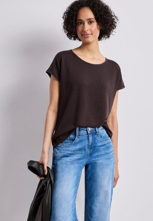 Kvinde med krøllet hår iført en løs mørkebrun top og lyseblå jeans, der holder en sort læderjakke i venstre hånd.