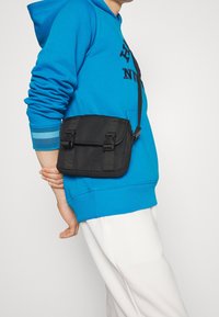 Borsa a tracolla nera con finitura testurizzata, dotata di chiusure a doppio pattina e tracolla regolabile, indossata insieme a un felpa blu brillante.